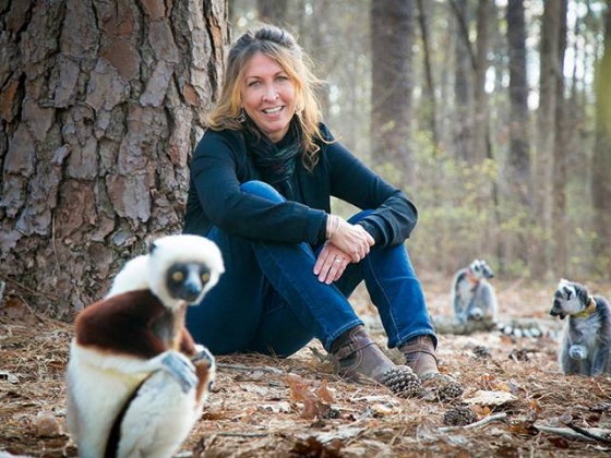 Anne Yoder smiles at the camera. She is sitting on the floor in front of a tree. Next to her are two ring-tailed lemurs. In first plan is a sifaka. 
