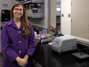 Elaine Guevara sales at the camera. She is leaning against a lab bench, next to a centrifuge and a set of pipettes.