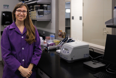 Elaine Guevara sales at the camera. She is leaning against a lab bench, next to a centrifuge and a set of pipettes.